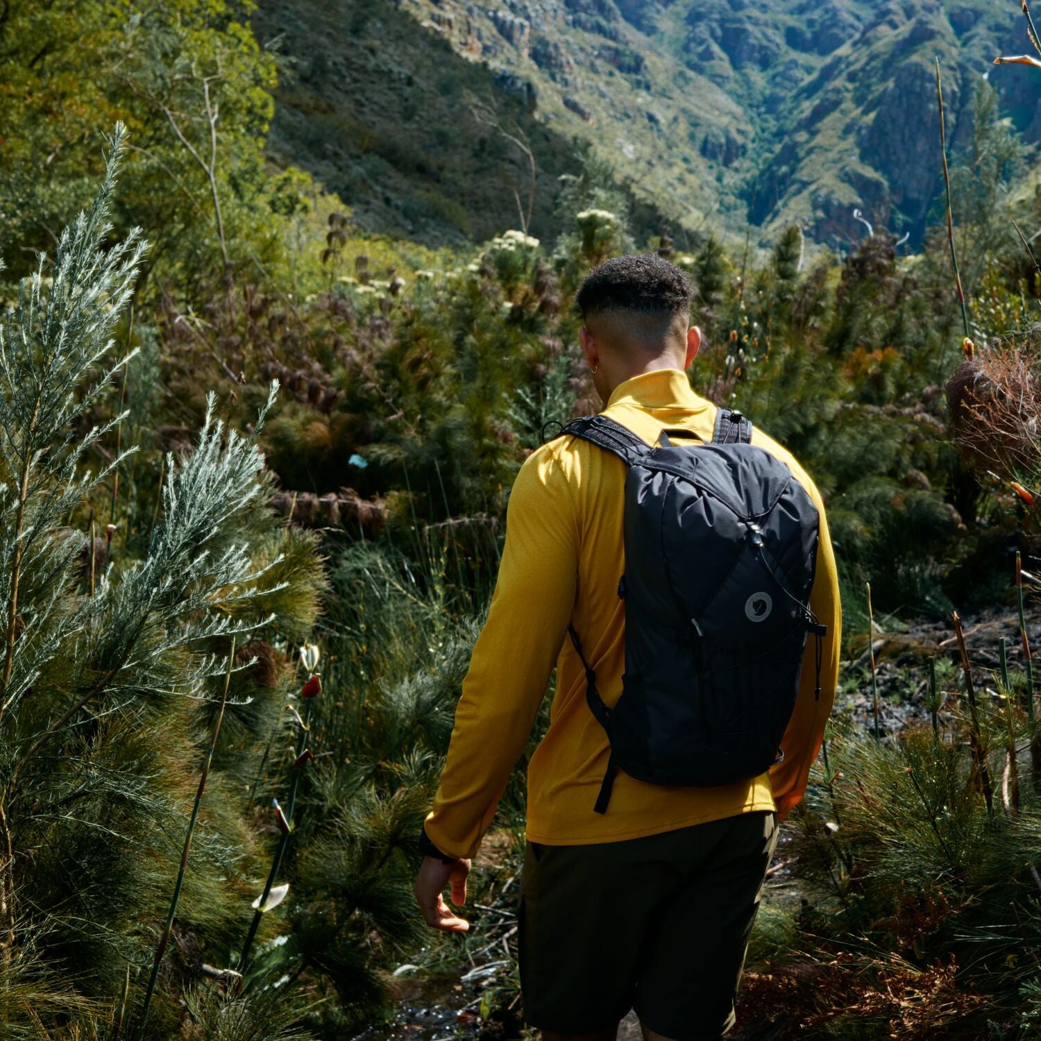Person in a yellow jacket with a black backpack walking through a forested area.
