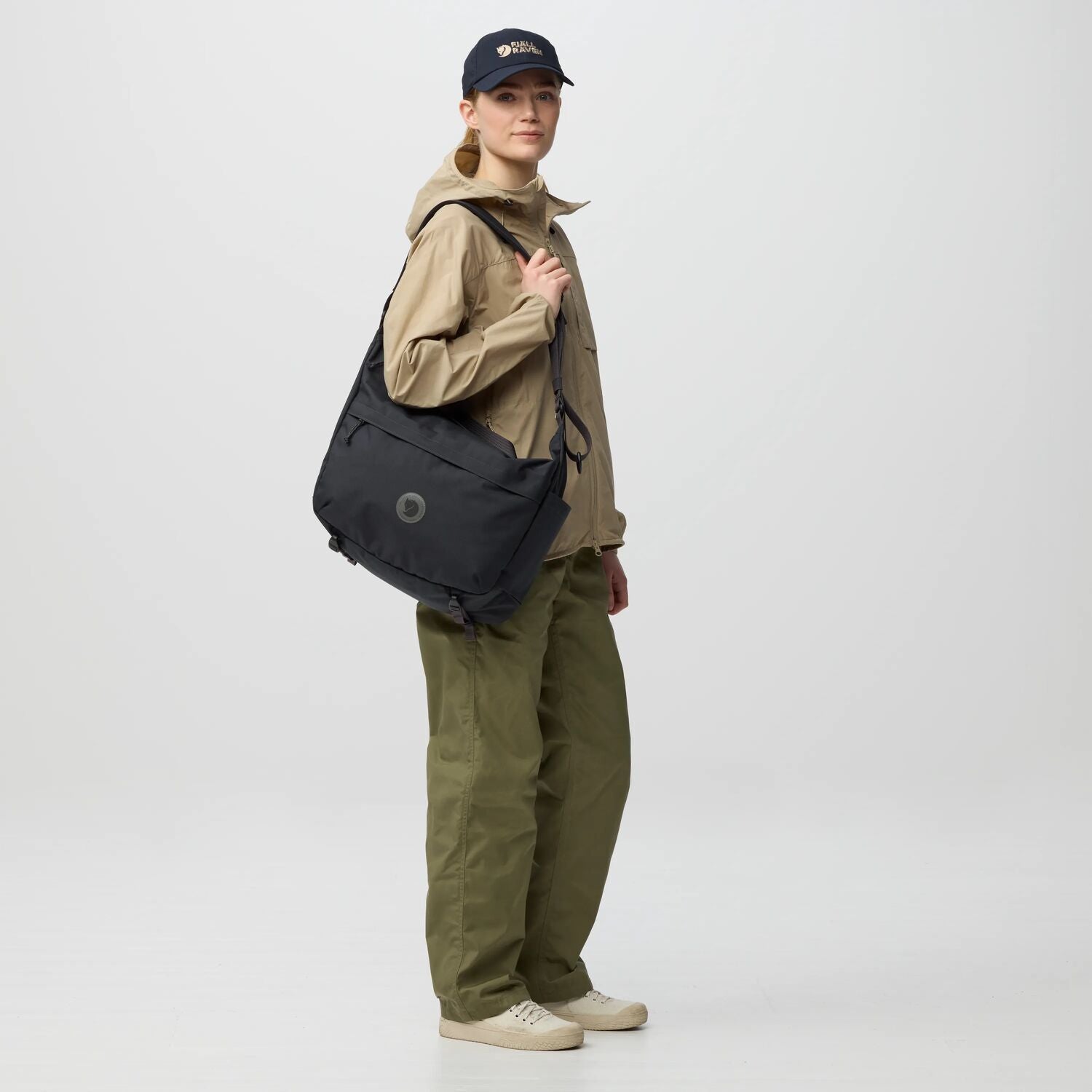 Person wearing a beige jacket, green pants, and a navy cap, holding a black bag on a white background