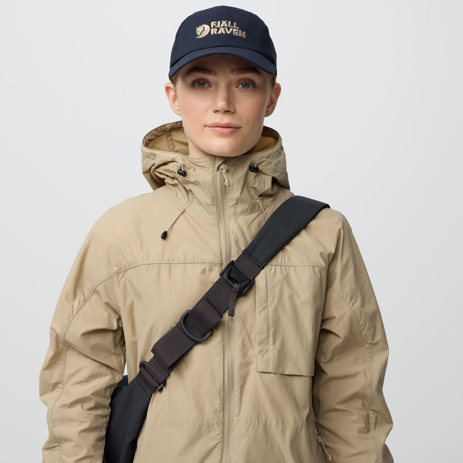 Person wearing a beige jacket and navy cap with 'Fjällräven' logo on a light gray background