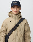 Person wearing a beige jacket and navy cap with 'Fjällräven' logo on a light gray background