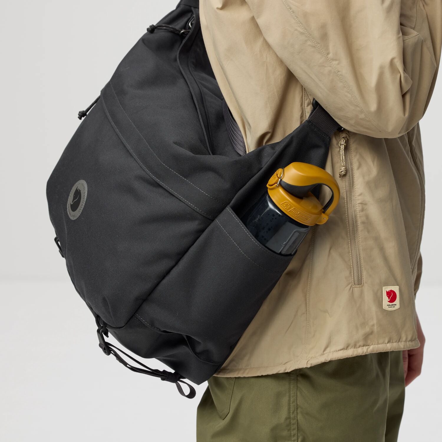 Person wearing a black backpack with a yellow handle, holding a beige jacket with a visible fjallraven logo.