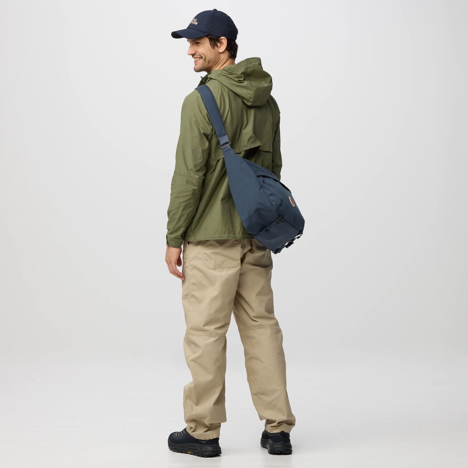 Man wearing a green jacket, beige pants, and a navy cap with a navy backpack on a white background