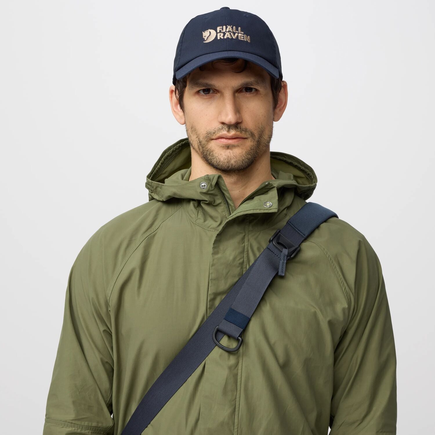 Man wearing a green raincoat and navy cap with a fjallraven logo on a light gray background