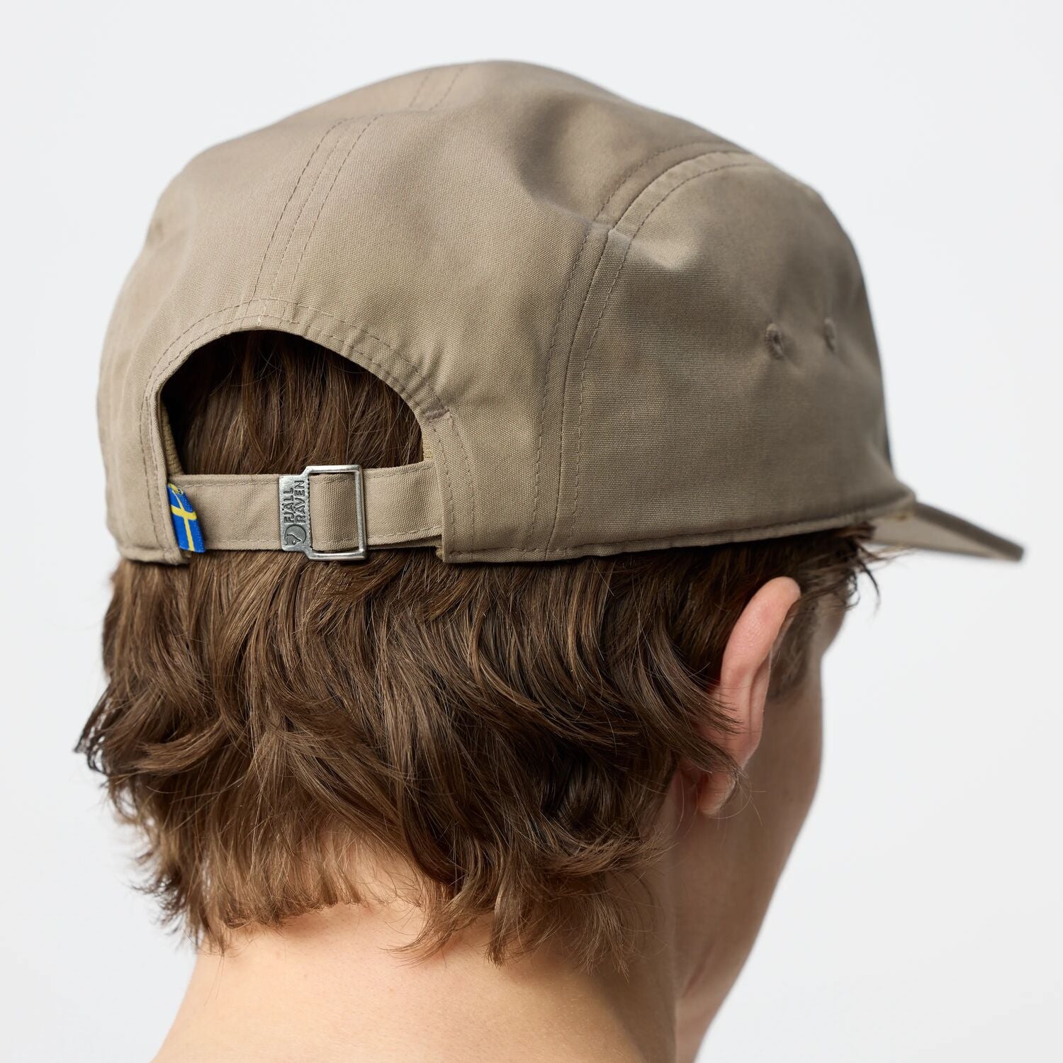 Beige cap with a small flag emblem on the back, worn by a person with short brown hair.
