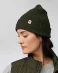 Person wearing a green beanie with a Fjällräven logo on a light gray background