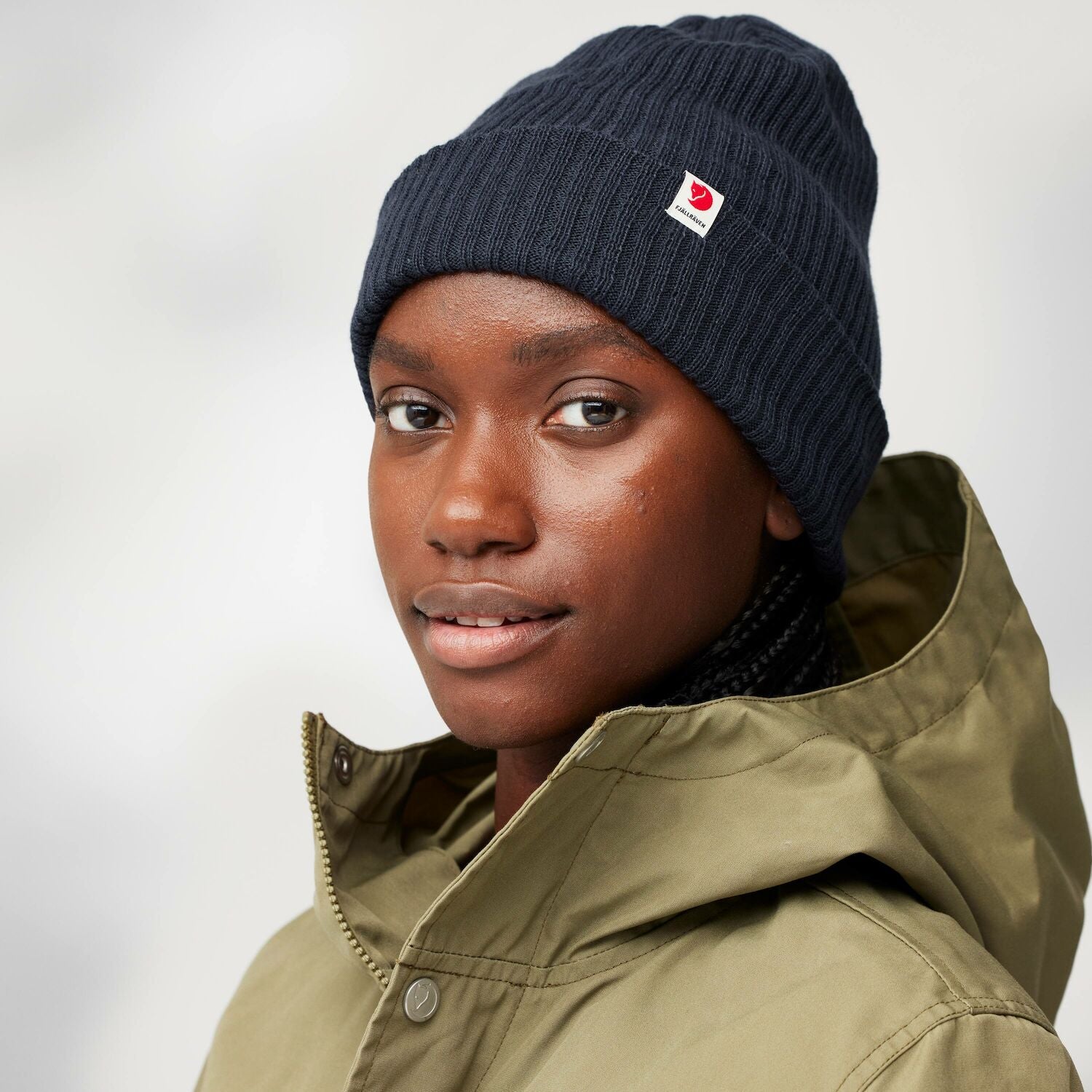 Person wearing a navy beanie with a Fjällräven logo and a green jacket on a light gray background