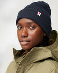Person wearing a navy beanie with a Fjällräven logo and a green jacket on a light gray background