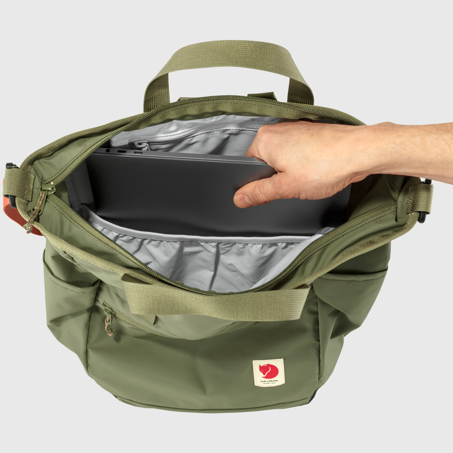 Green backpack with a hand reaching inside to place a black item, on a light gray background