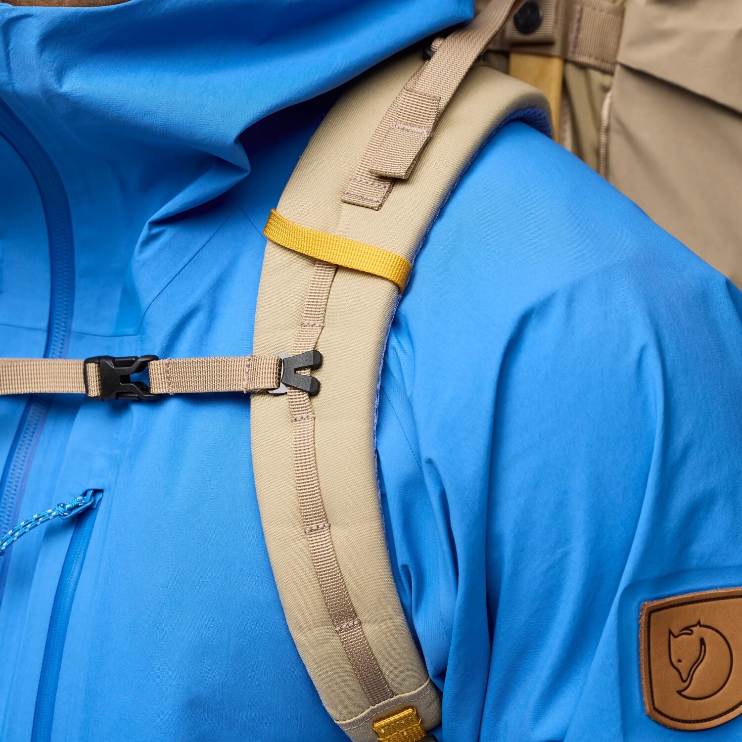 Close-up of a blue jacket with a beige strap and a brand logo on a person wearing it.