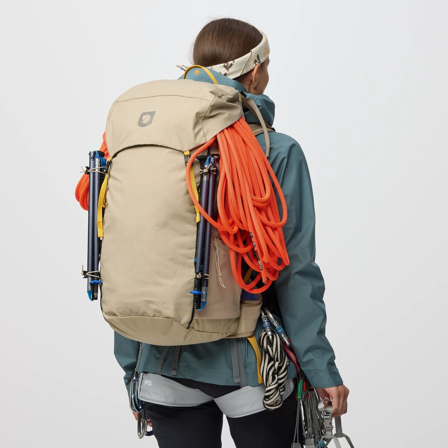 Person wearing a large beige backpack with climbing equipment on a plain background