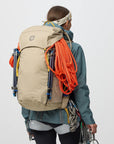 Person wearing a large beige backpack with climbing equipment on a plain background