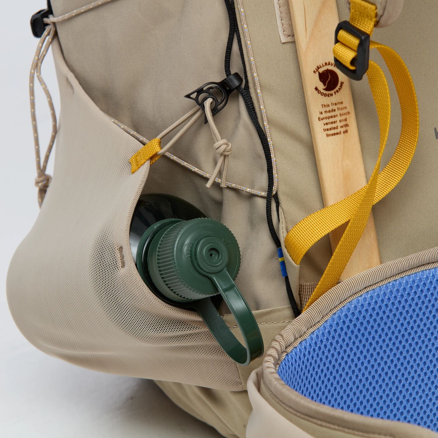 Close-up of a beige backpack with a green water bottle and wooden stability structure, on a white background.