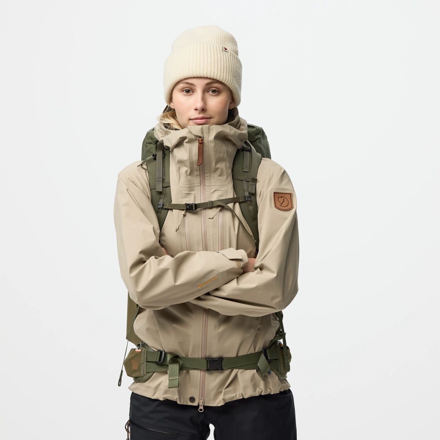 Person wearing a beige jacket, black pants, and a white beanie with a green backpack on a light gray background