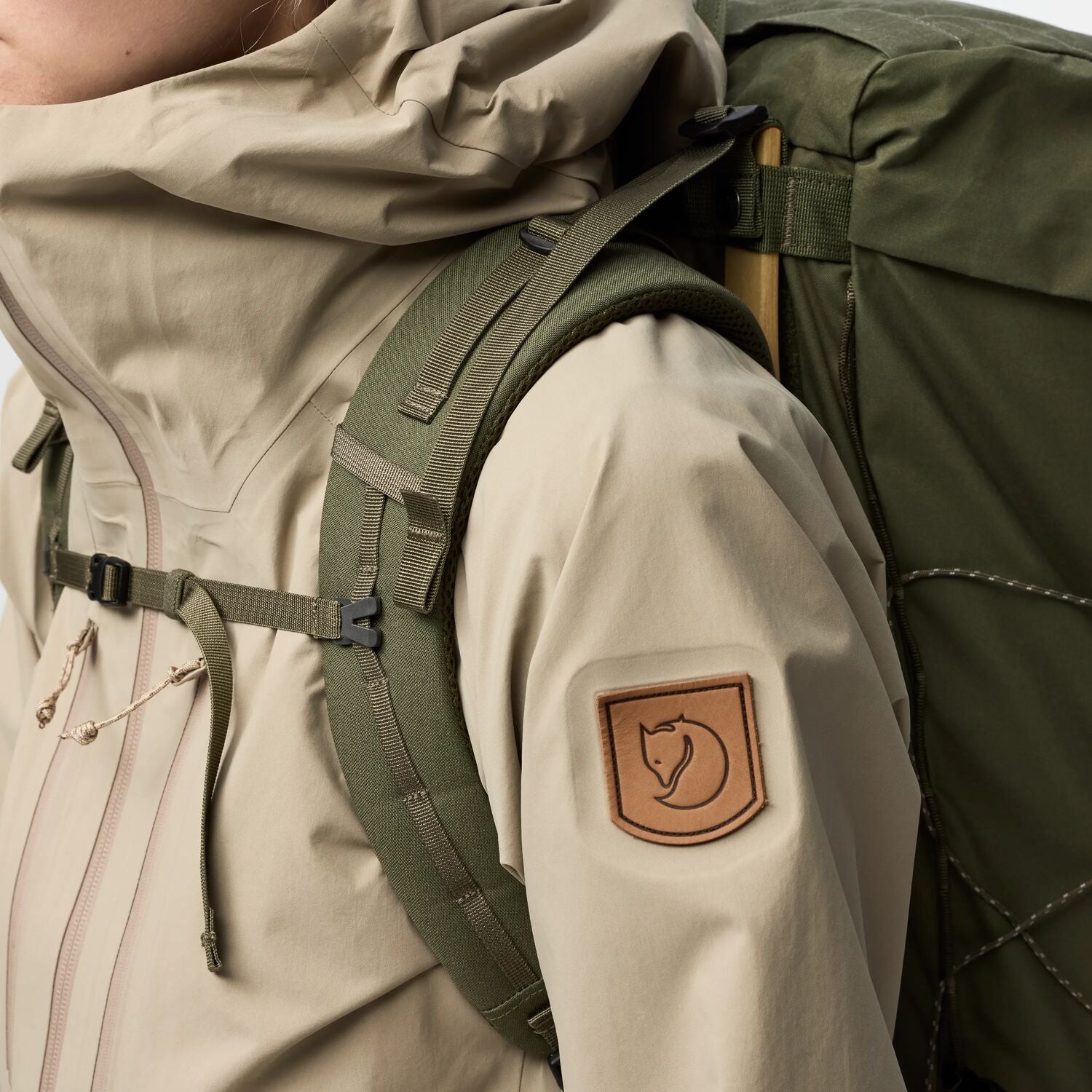 Person wearing a beige jacket with a green backpack, featuring a brand logo on the sleeve.