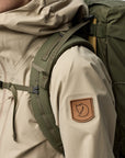 Person wearing a beige jacket with a green backpack, featuring a brand logo on the sleeve.
