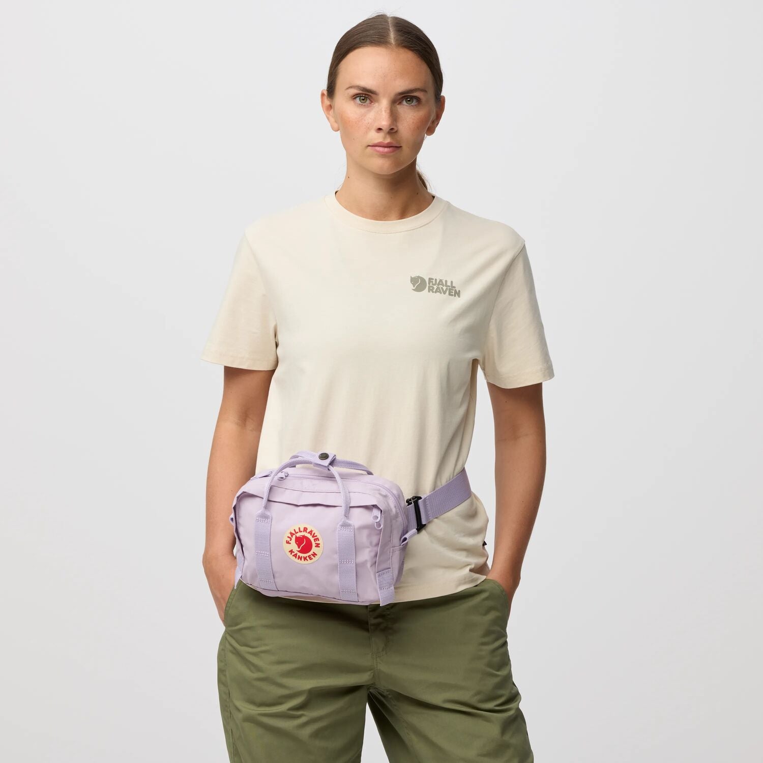 Person wearing a beige t-shirt and green pants with a purple waist bag on a light gray background
