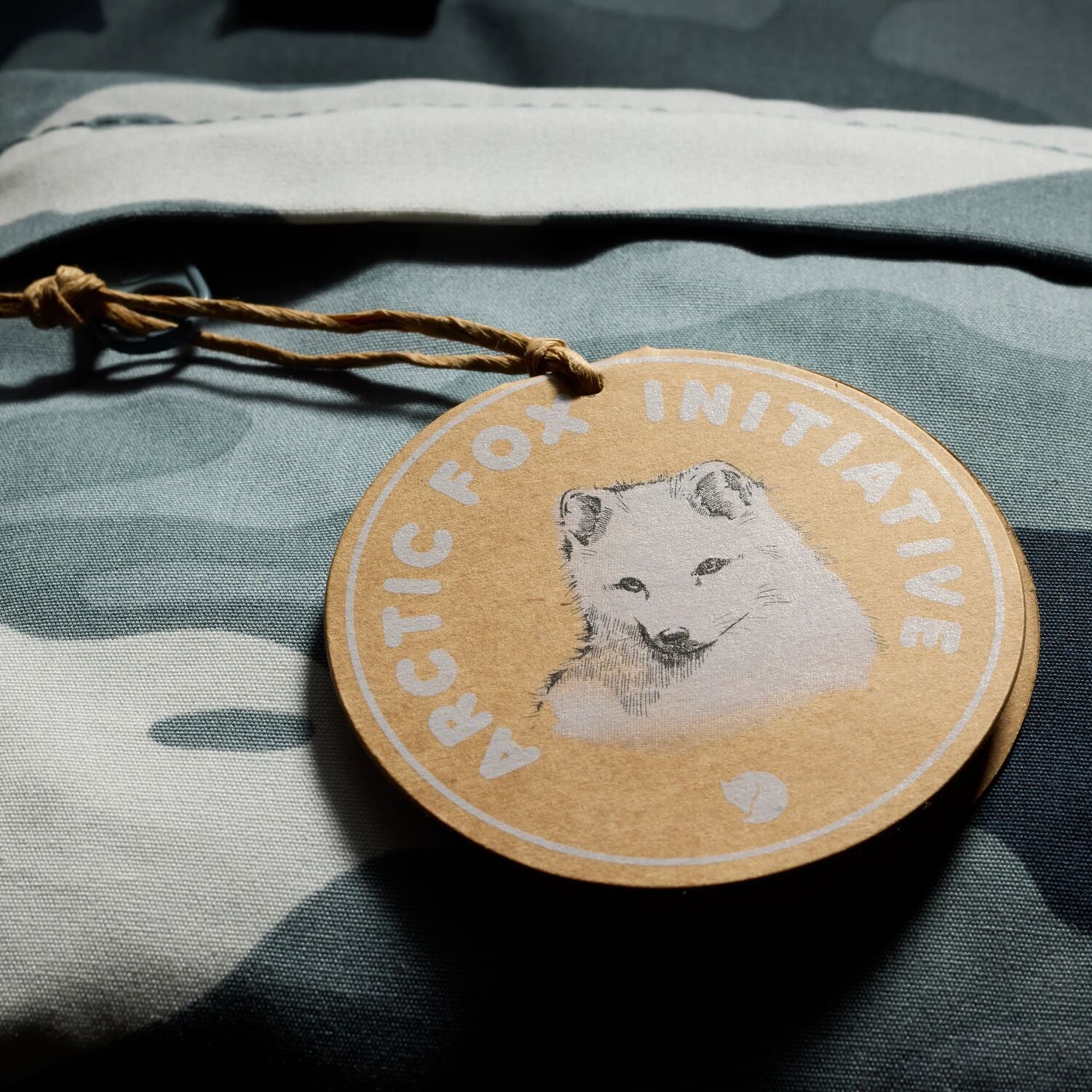 Round wooden tag with an arctic fox illustration and 'Arctic Fox Initiative' text on a camouflage fabric background.