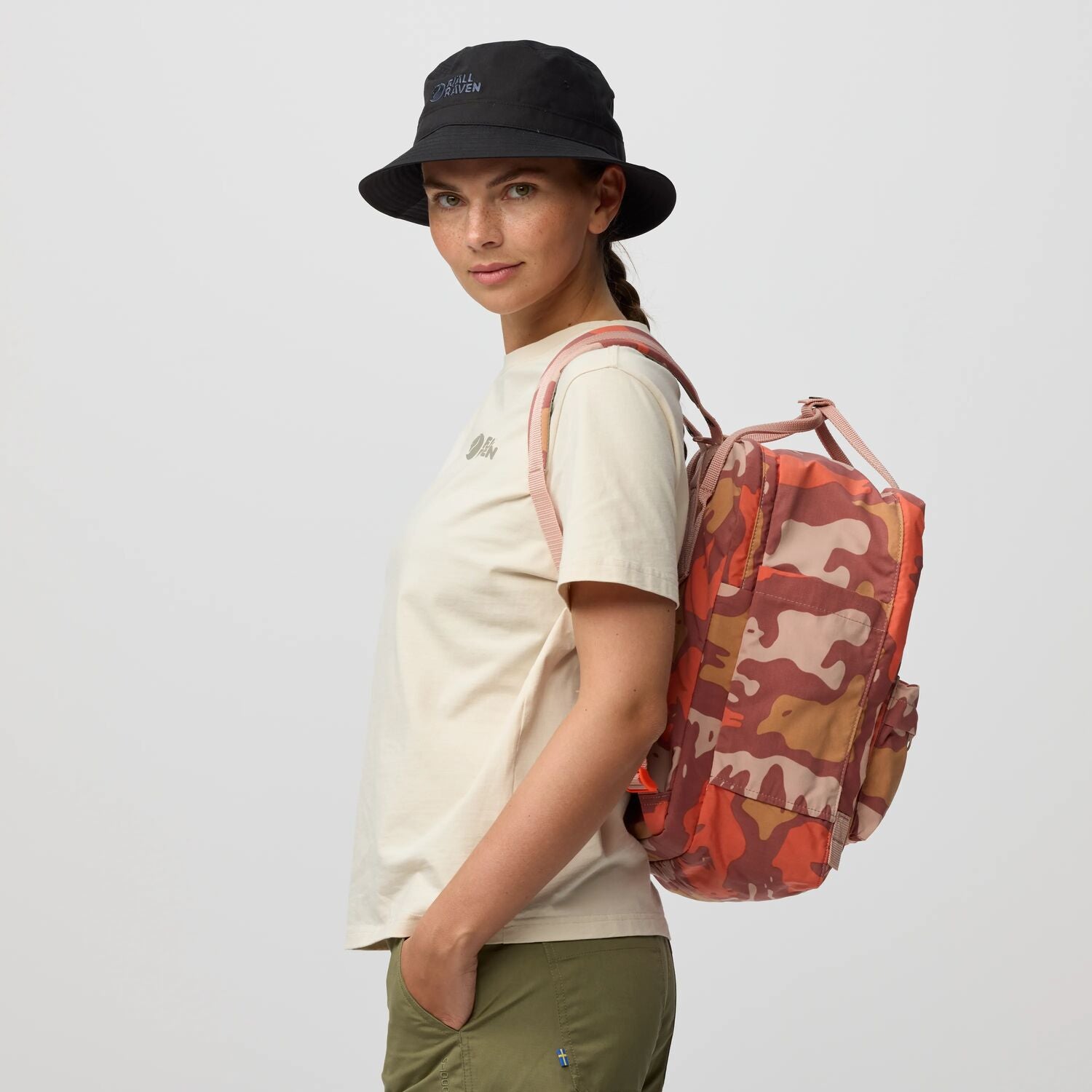 Person wearing a white t-shirt, black bucket hat, and carrying a red camouflage backpack on a light gray background