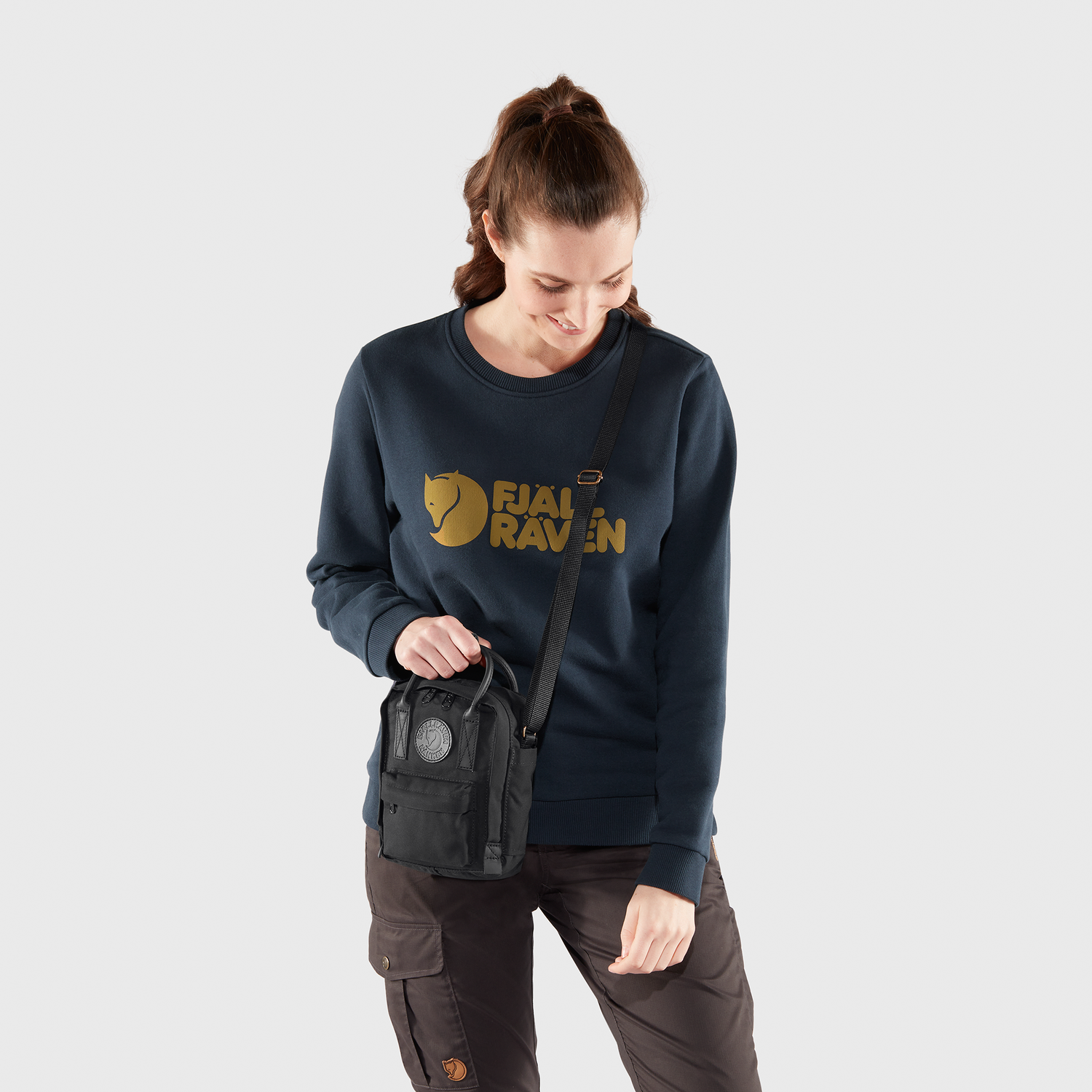 Woman wearing a navy Fjällräven sweatshirt holding a black bag on a light gray background