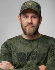 Man wearing a green cap and long-sleeve shirt with 'Fjällräven' logo on a gray background