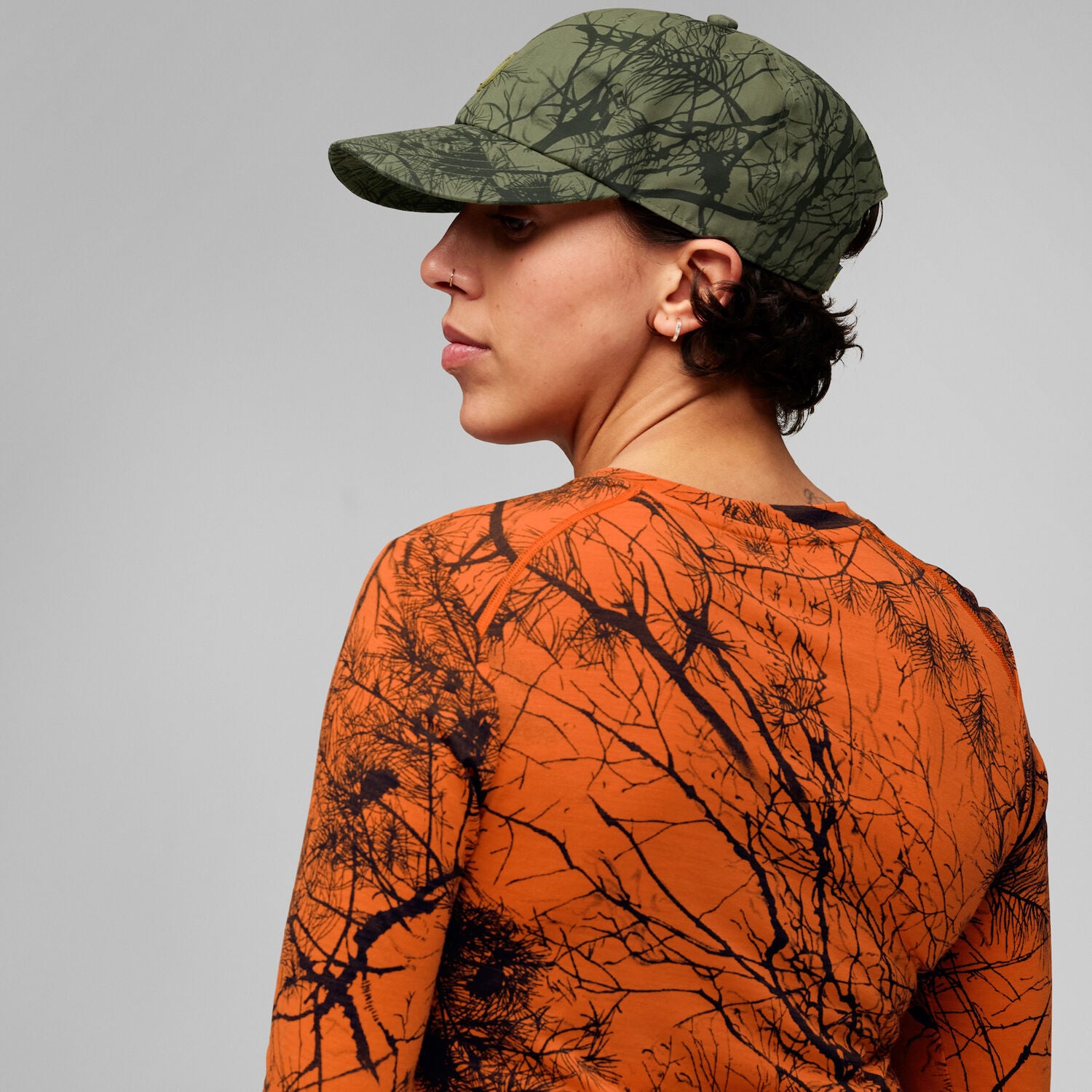 Person wearing a camouflage cap and shirt with a tree branch pattern on a gray background