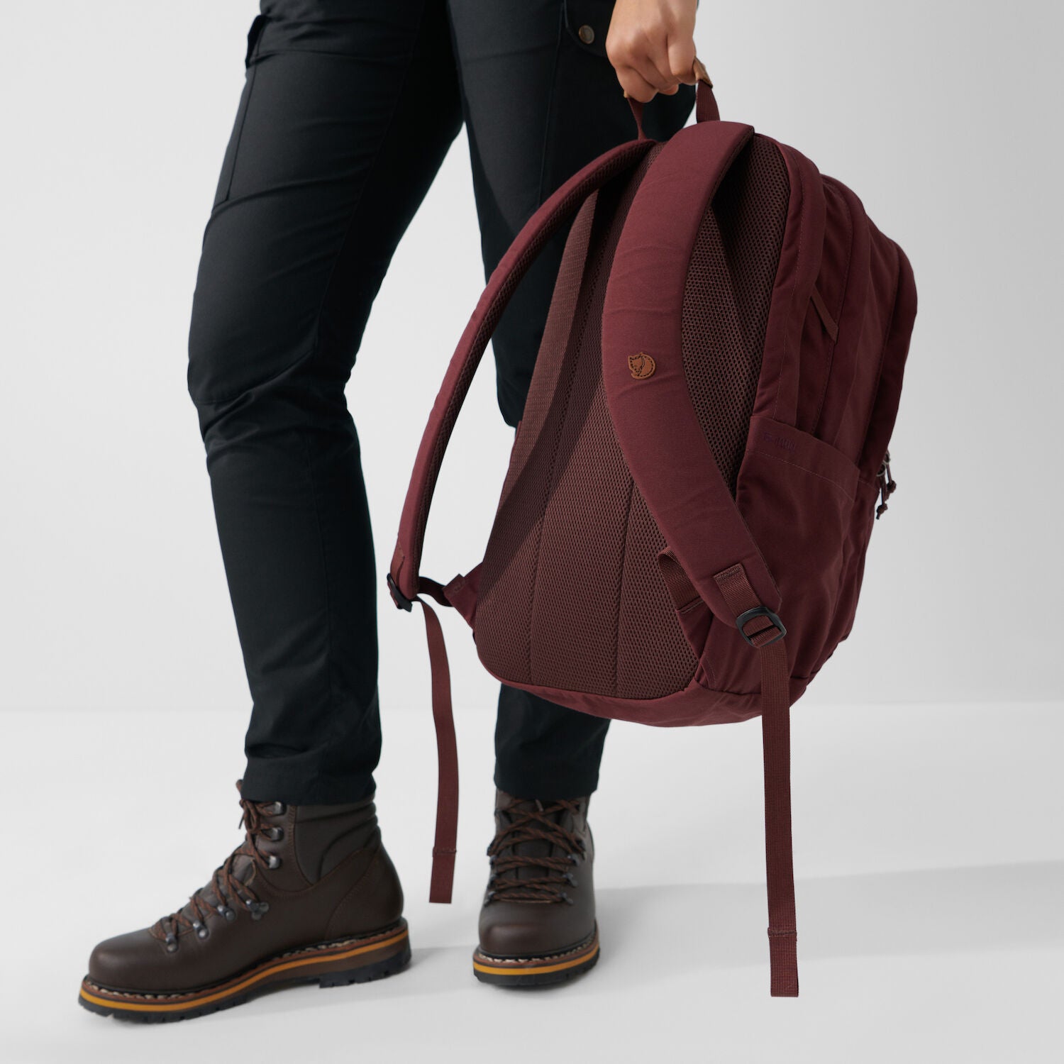 Person holding a maroon backpack with black pants and brown boots on a light gray background