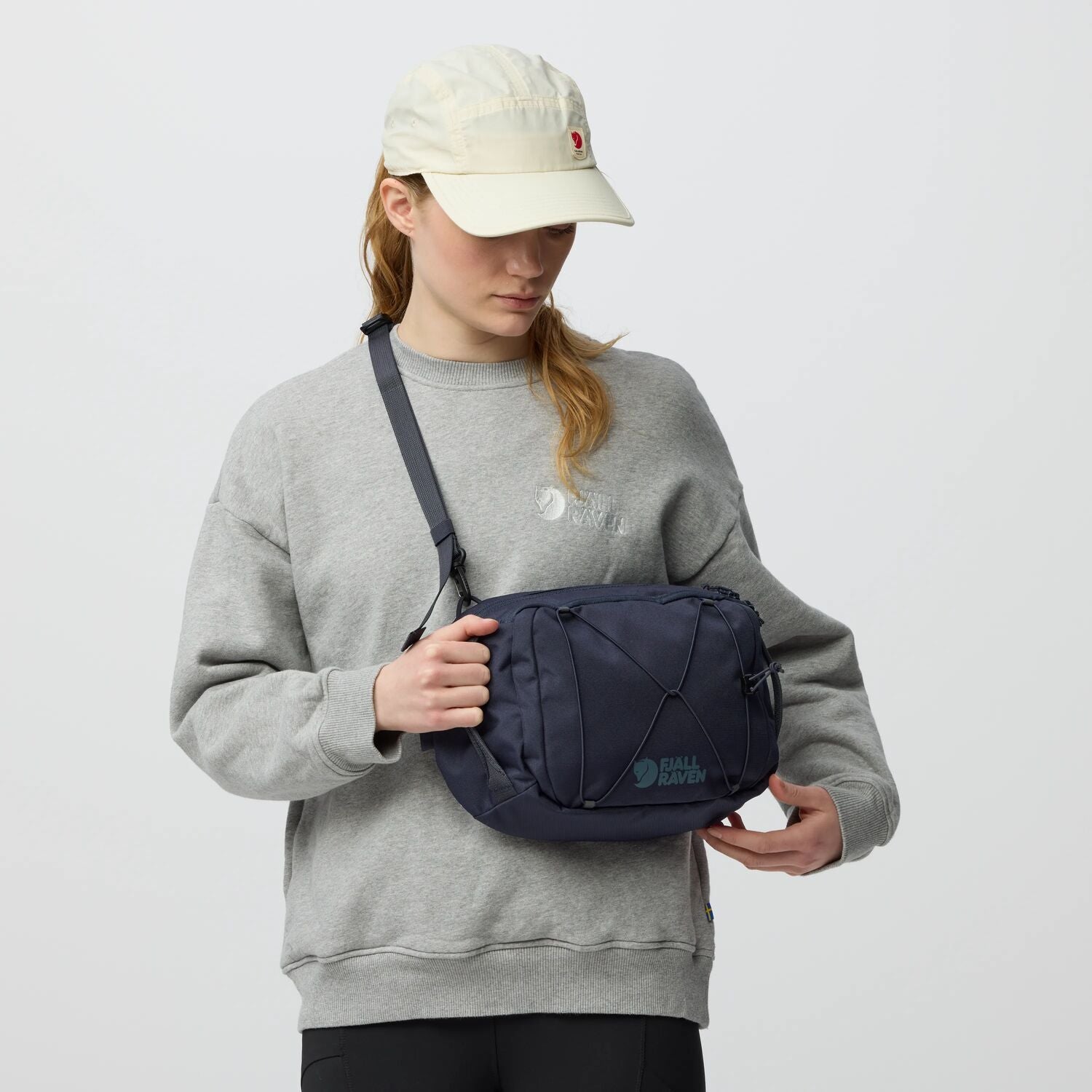 Person wearing a gray sweatshirt and beige cap holding a navy blue bag against a light gray background