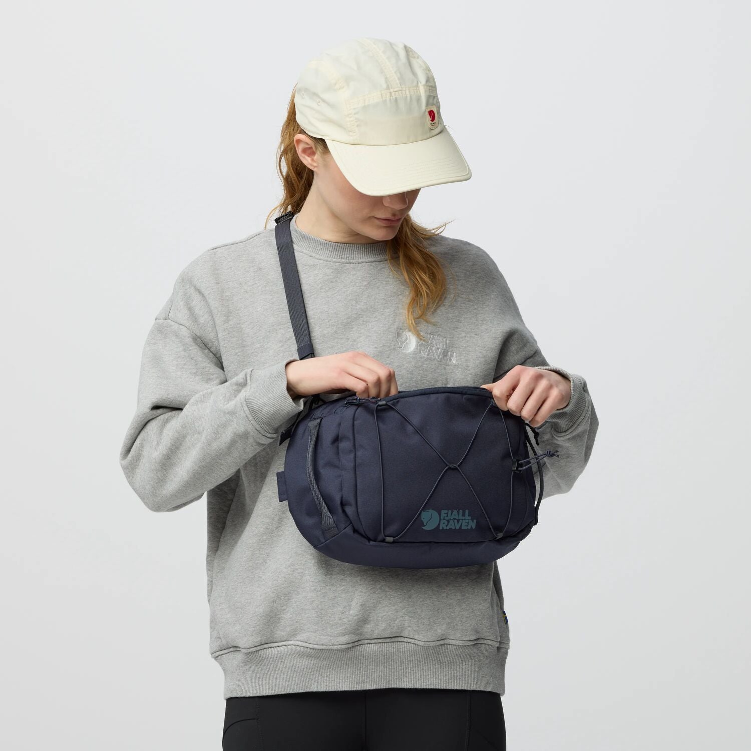 Person wearing a gray sweatshirt and beige cap holding a navy blue bag against a white background