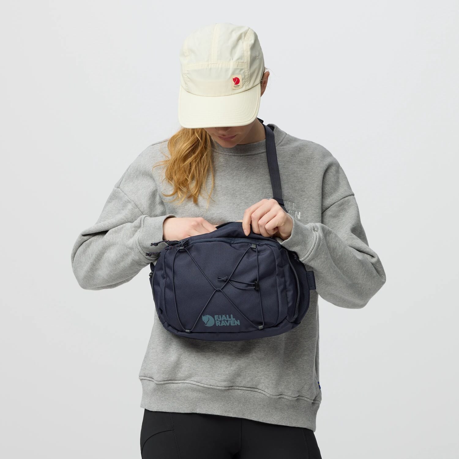 Person wearing a gray sweatshirt and white cap, holding a navy blue bag with a visible brand logo.