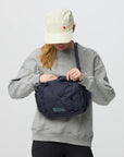 Person wearing a gray sweatshirt and white cap, holding a navy blue bag with a visible brand logo.