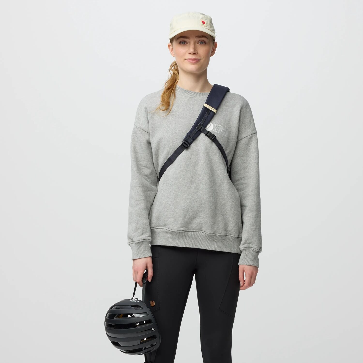 Person wearing a gray sweatshirt and black pants, holding a helmet, on a light gray background