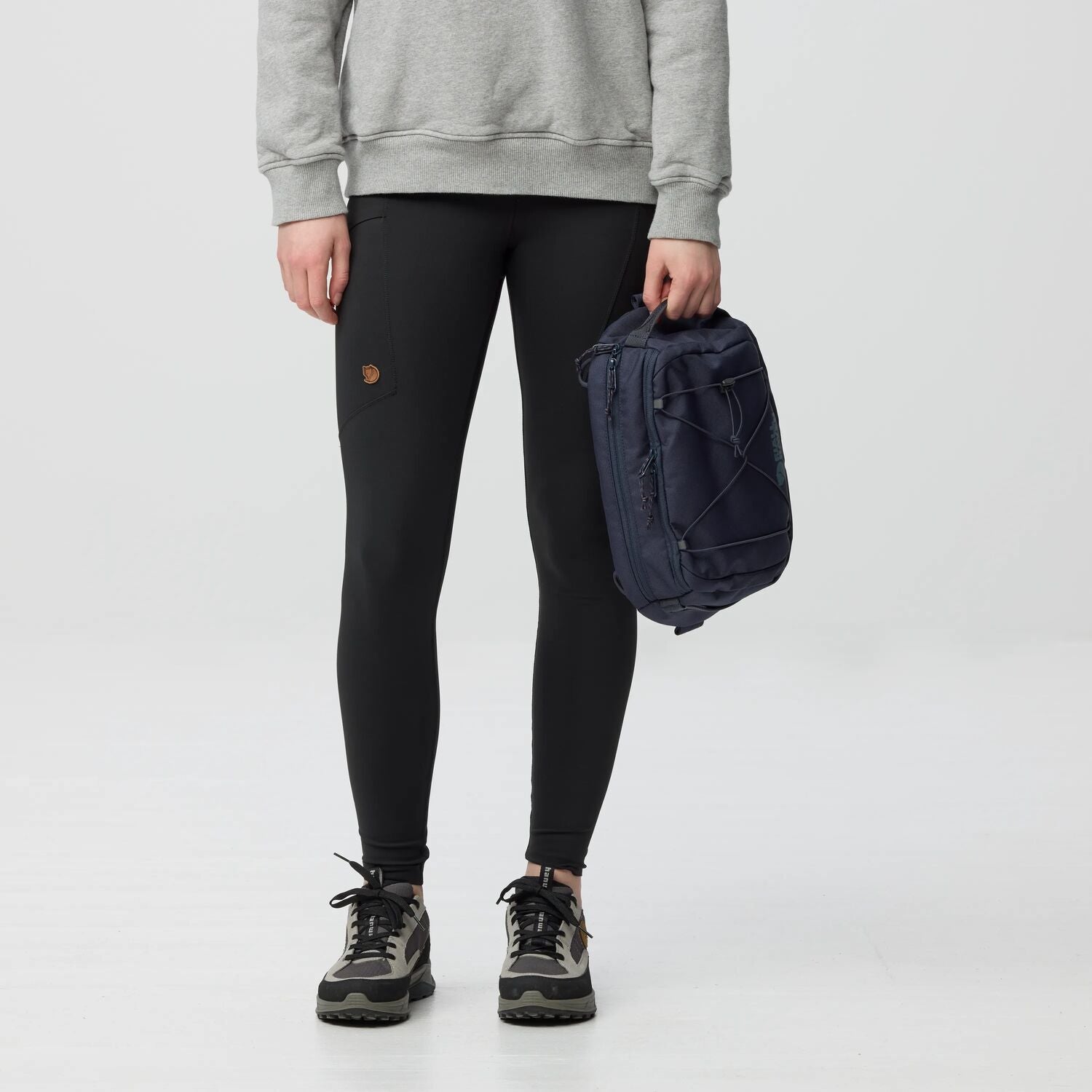 Person wearing black leggings with a logo, holding a navy backpack on a light gray background