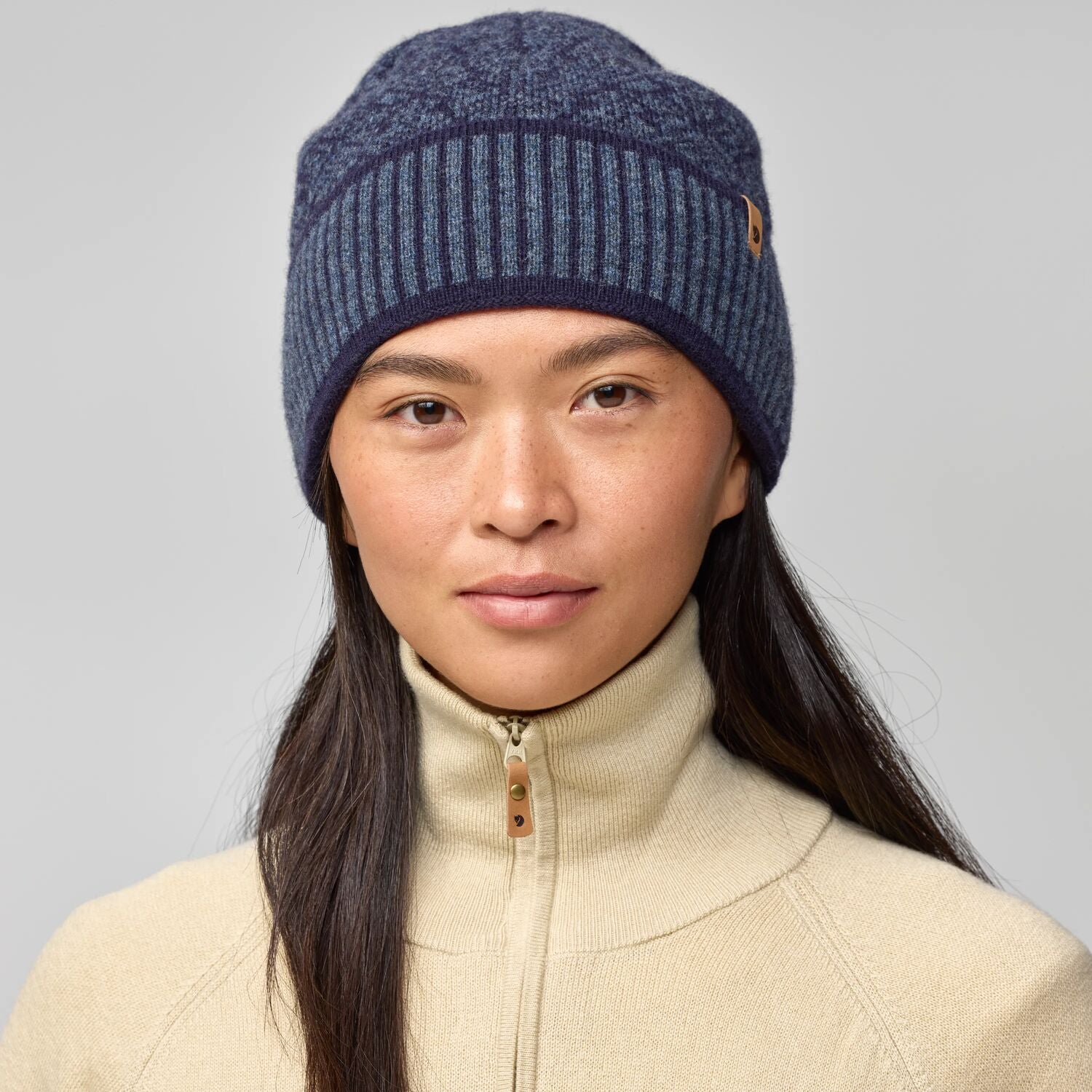 Woman wearing a blue knit beanie and beige jacket on a gray background
