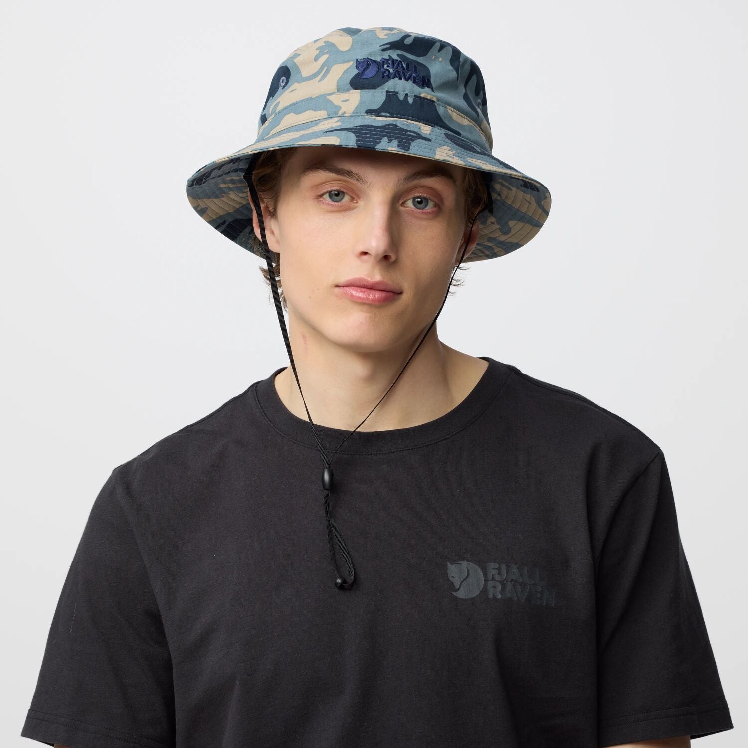 Person wearing a camouflage bucket hat and black t-shirt with a Fjällräven logo on a light gray background