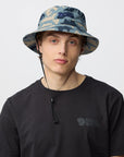 Person wearing a camouflage bucket hat and black t-shirt with a Fjällräven logo on a light gray background