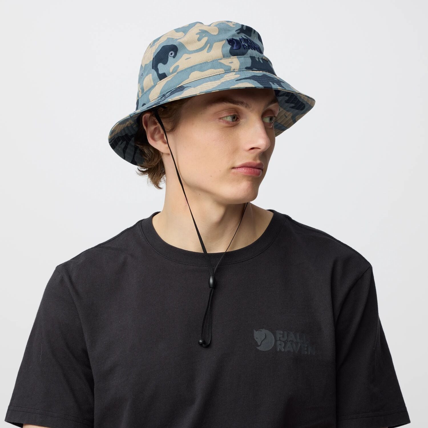 Person wearing a camouflage bucket hat and black t-shirt with a Fjällräven logo on a light gray background