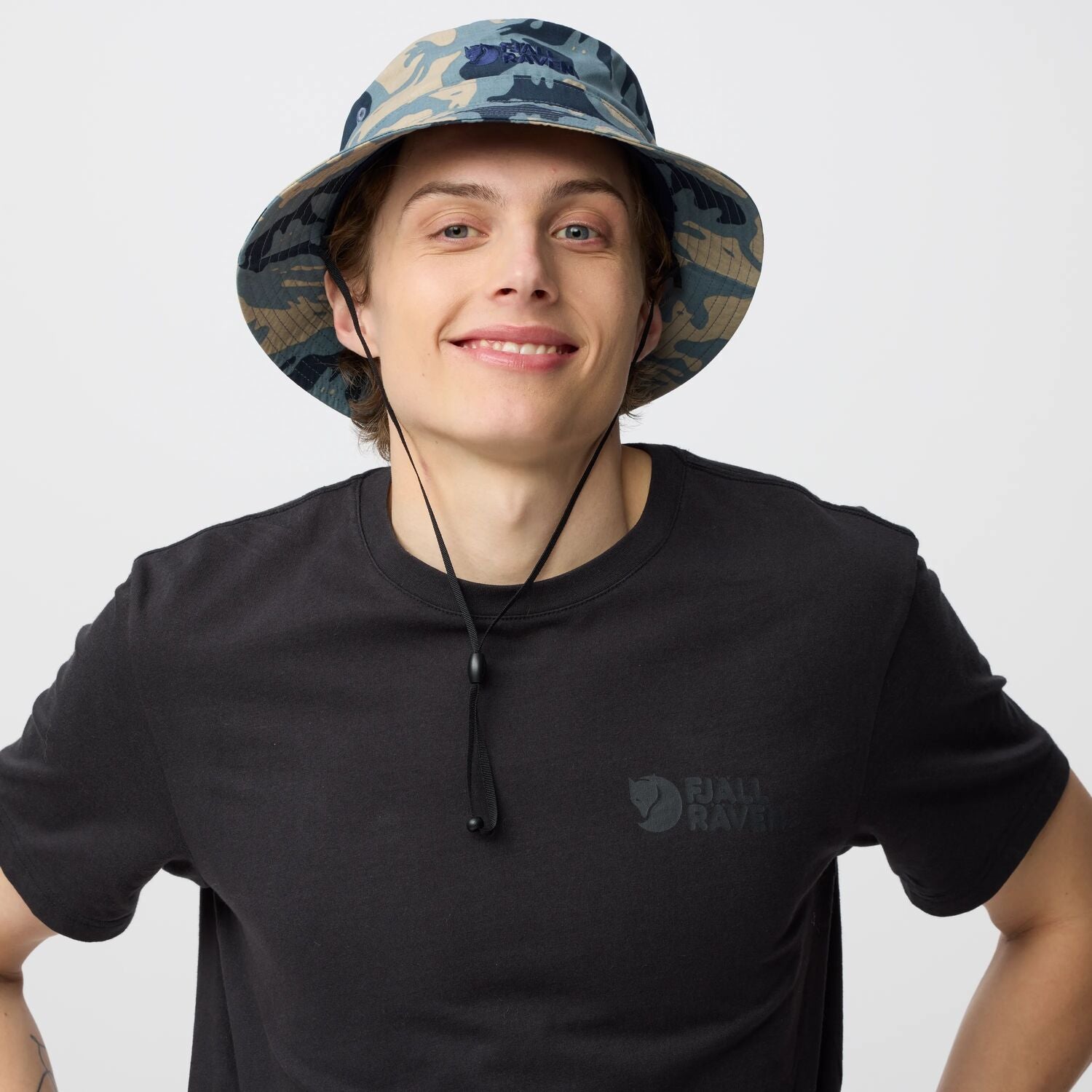 Person wearing a black t-shirt with a  Fjällräven logo and a camouflage bucket hat on a light gray background