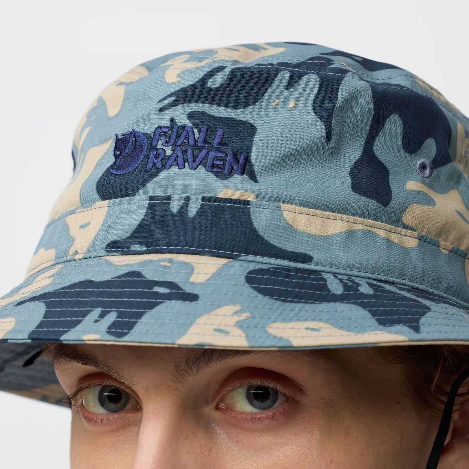 Person wearing a camouflage bucket hat with Fjällräven logo on a white background