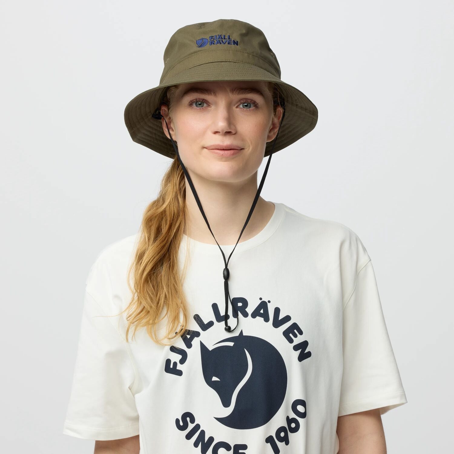 Person wearing a white t-shirt with Fjälåråven logo and a green bucket hat on a light gray background