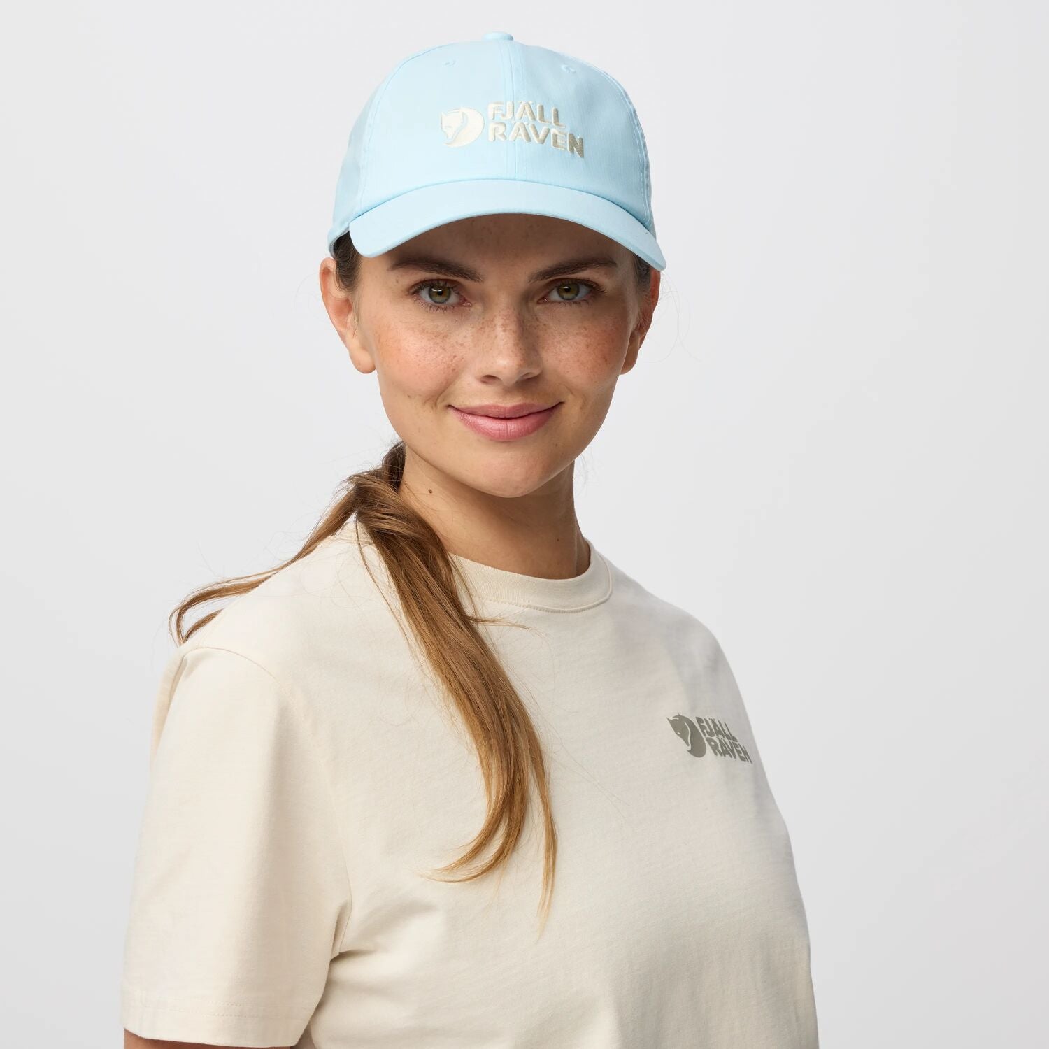 Woman wearing a light blue cap and beige t-shirt with a logo on a plain background
