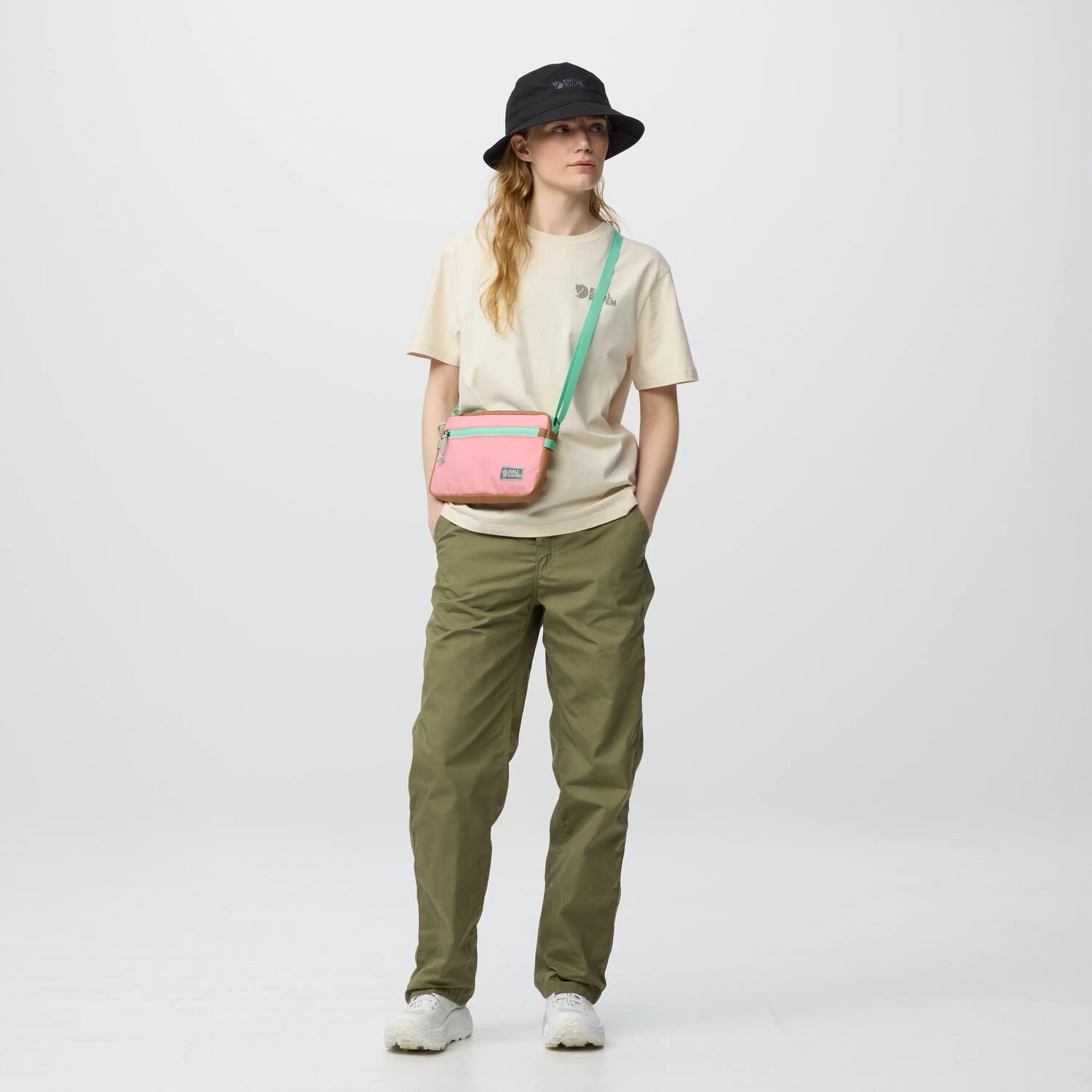 Person wearing a beige shirt, green pants, and a black hat, carrying a pink crossbody bag on a white background.