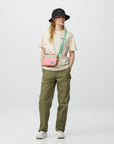 Person wearing a beige shirt, green pants, and a black hat, carrying a pink crossbody bag on a white background.