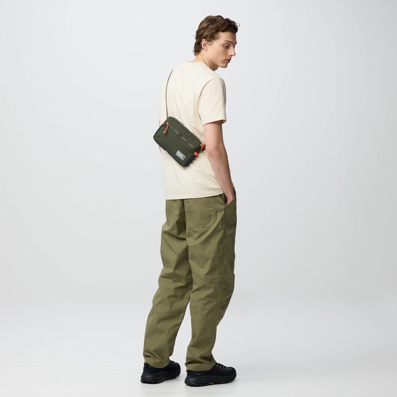 Person wearing a beige shirt and green pants with a green bag on a white background