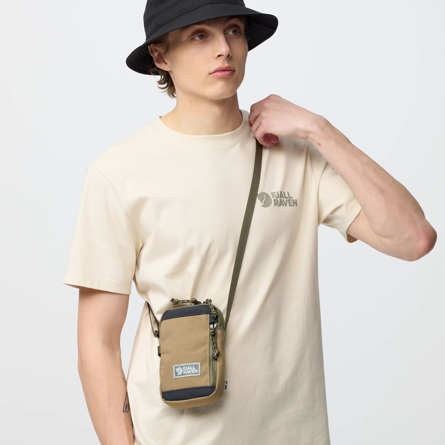 Person wearing a beige t-shirt with a logo and carrying a small bag on a plain background