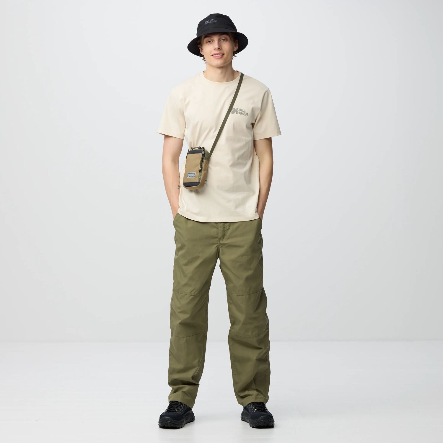 Person wearing a beige t-shirt, green pants, black shoes, and a black hat on a white background