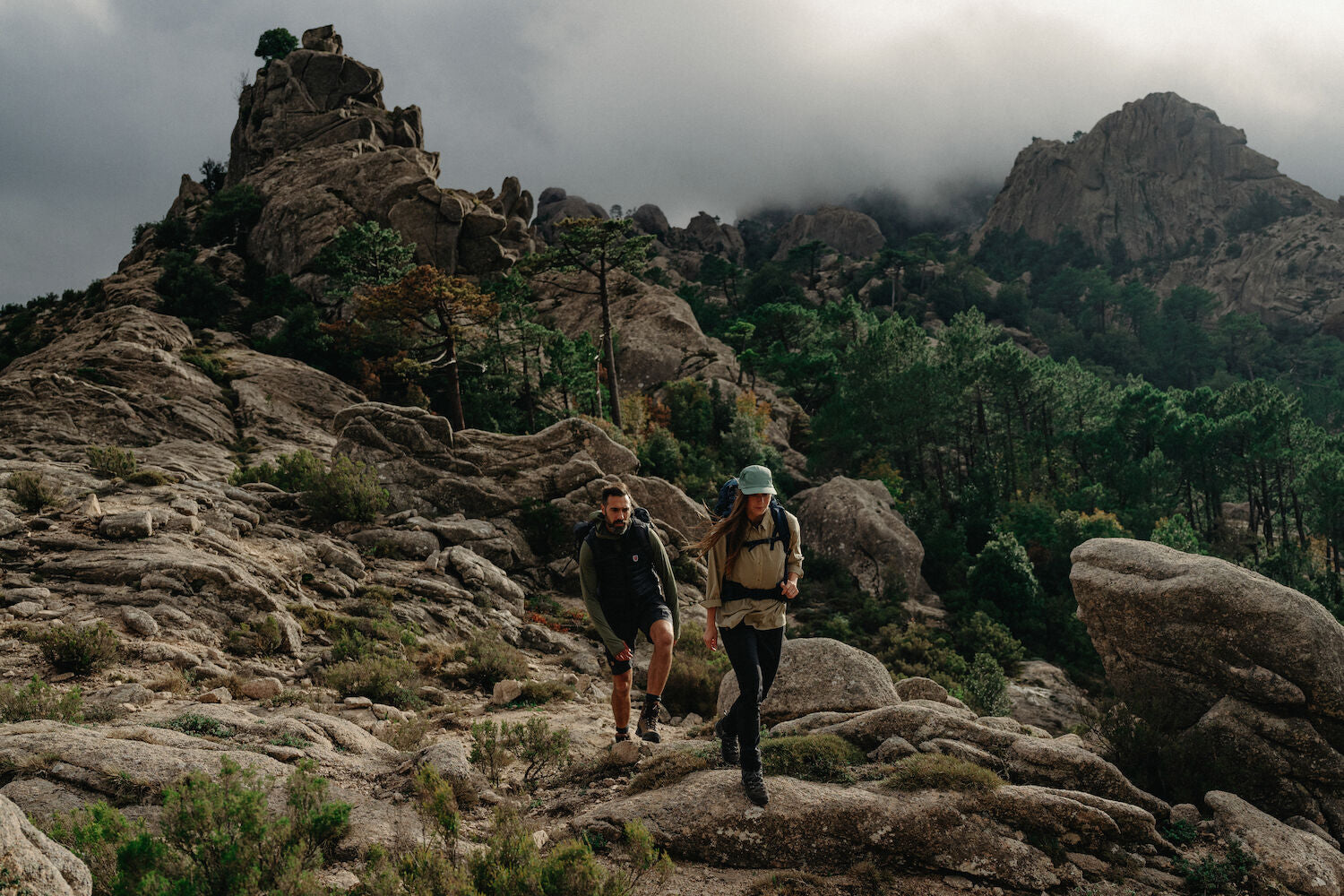 Two hikers on a rocky mountain trail with a cloudy sky.