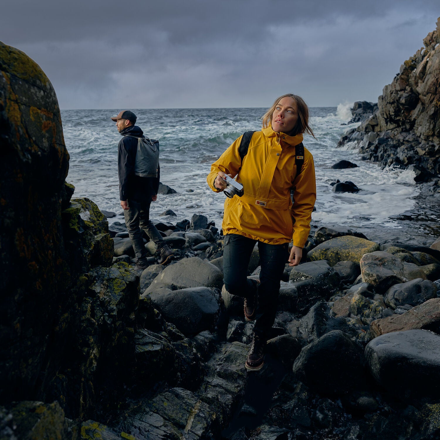 woman wearing mustard yellow waterproof rain jacket with Fjällräven logo walking on rocky coast