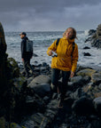 woman wearing mustard yellow waterproof rain jacket with Fjällräven logo walking on rocky coast