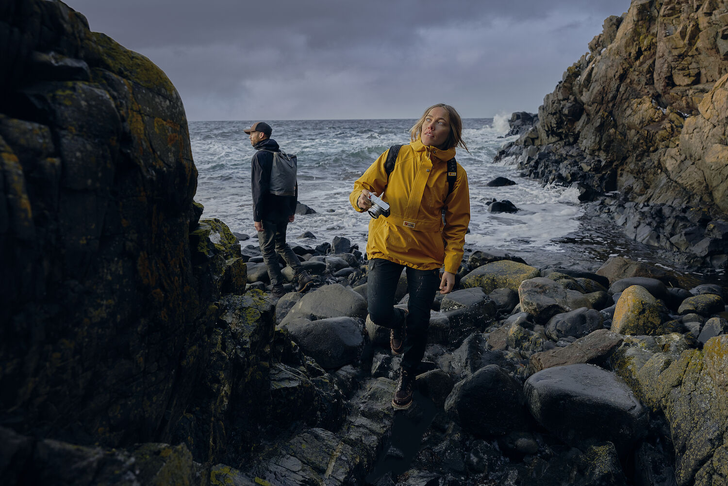 woman wearing yellow raincoat jacket with Fjällräven logo holding a camera and walking on stone surface