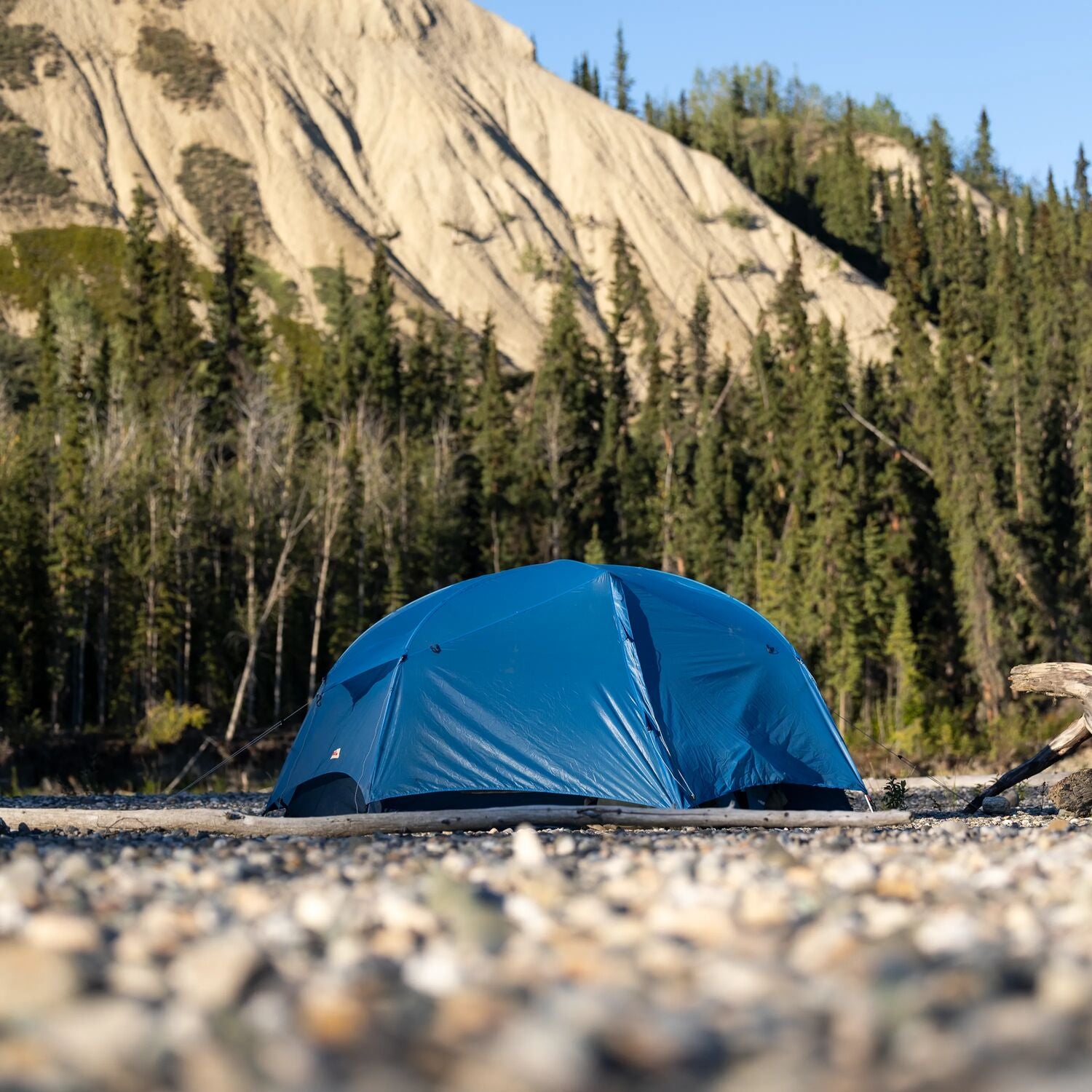 Blue tent set up on a rocky ground with trees and mountains in the background